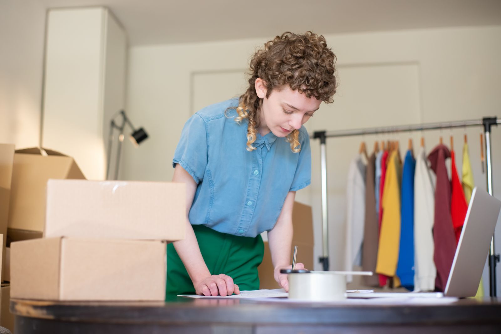 A small-brand founder taping up an order in her home studio, with stacked boxes and a rack of inventory behind her