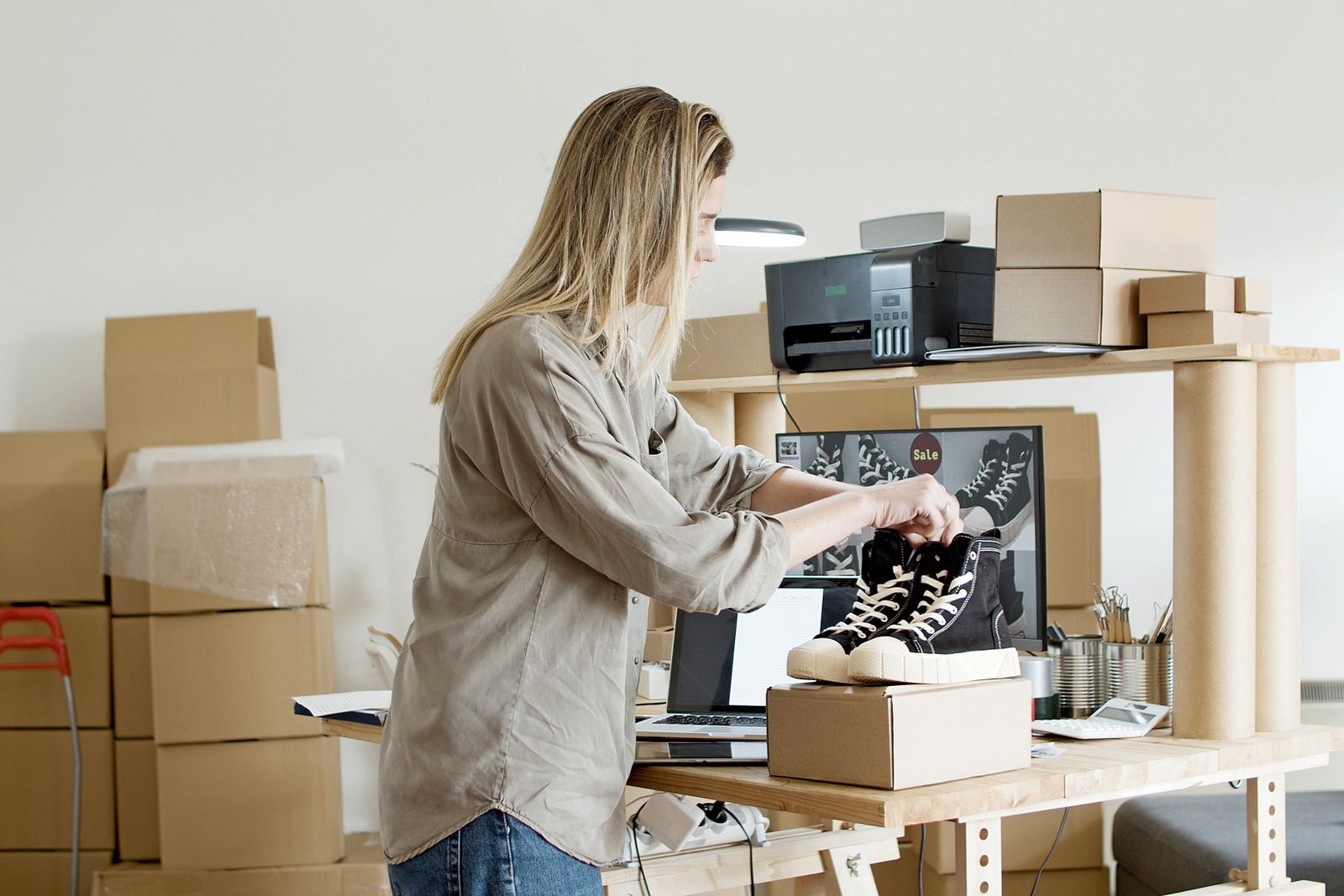 A small-brand founder packing an order at home, shelves of inventory and boxes behind her