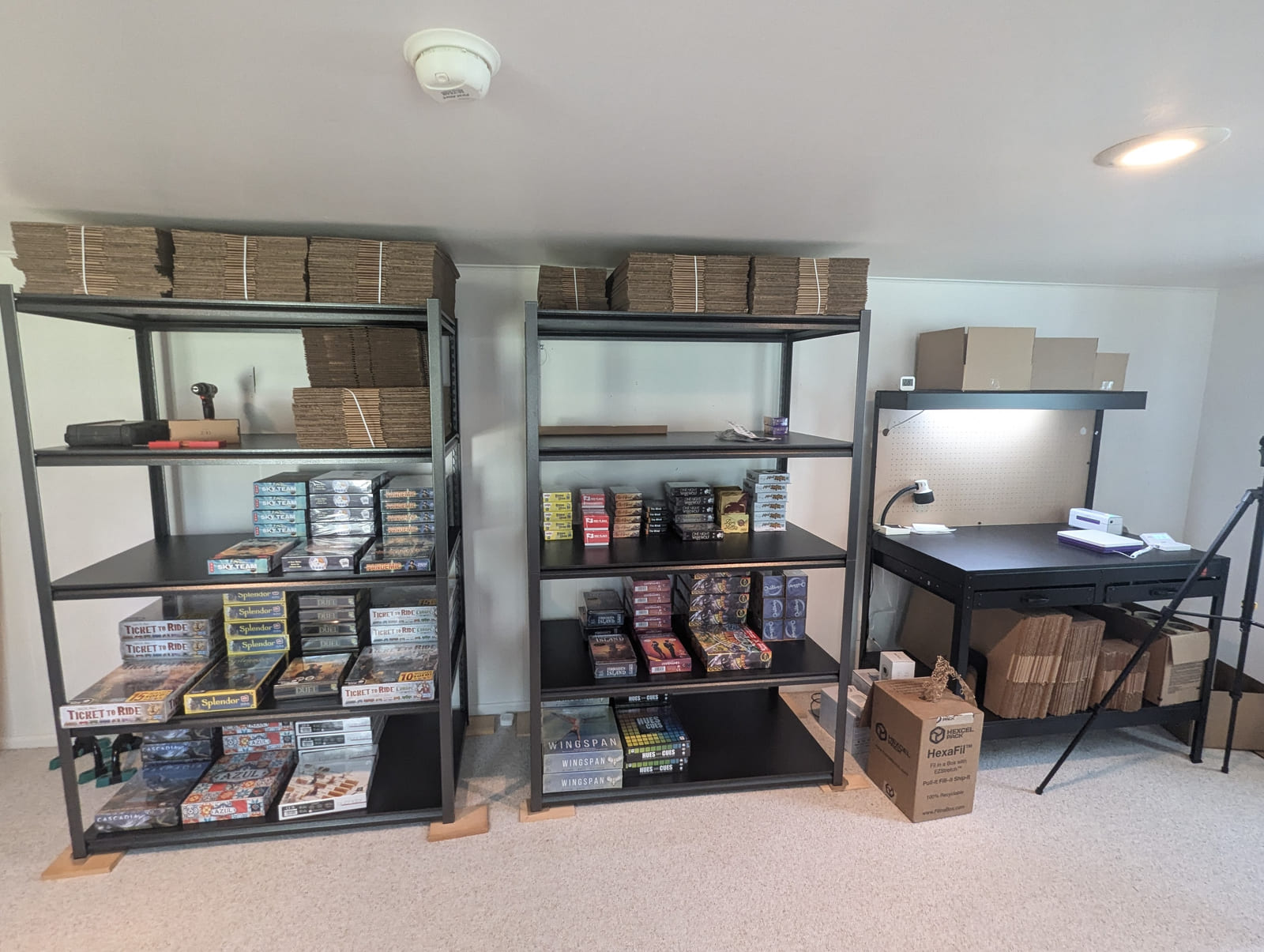 A home fulfillment room run by a Middle Mile operator: two steel shelving units stocked with boxed inventory, flat-packed cartons stored on top, and a packing bench with a label printer and task light.